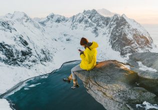 Woman sitting on cliff and using smartphone on Lofoten island in snow.