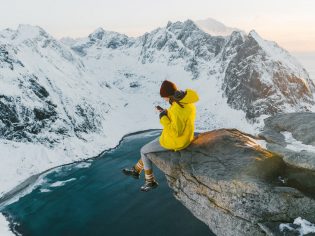 Woman sitting on cliff and using smartphone on Lofoten island in snow.