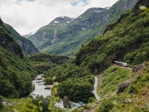Passenger train of the Flåm Railway Line passes along a mountainside with a glacial river in the valley below. The line runs between Myrdal and Flåm in Aurland