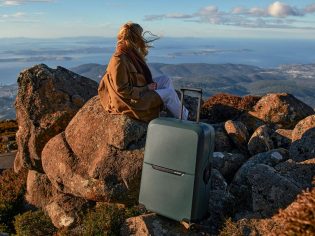 A woman with a brown jacket sitting on a rock with a grey suitcase.