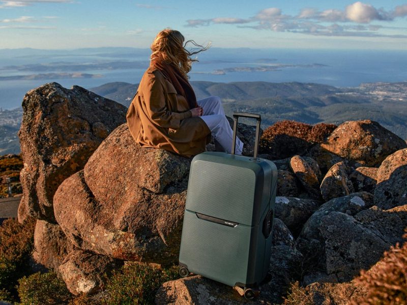 A woman with a brown jacket sitting on a rock with a grey suitcase.