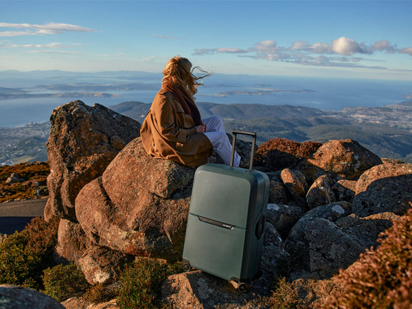 A woman with a brown jacket sitting on a rock with a grey suitcase.