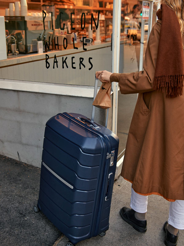 A woman with a brown jacket and scarf walking in the street with a blue suitcase.