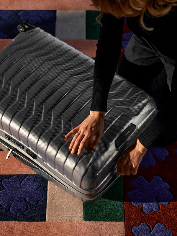 A woman opening a grey suitcase on a rug.