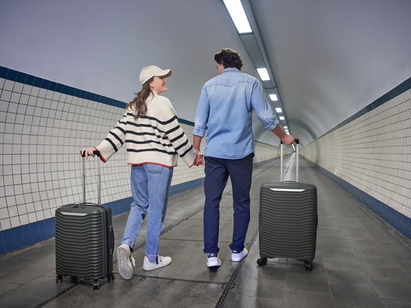 A woman with a white sweater and hat with jeans holding a grey suitcase and a man's hand who has a blue shirt and blue pants and is also holding a black suitcase.