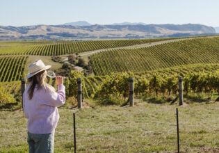 A woman sipping wine in front of a vineyard in Marlborough New Zealand.