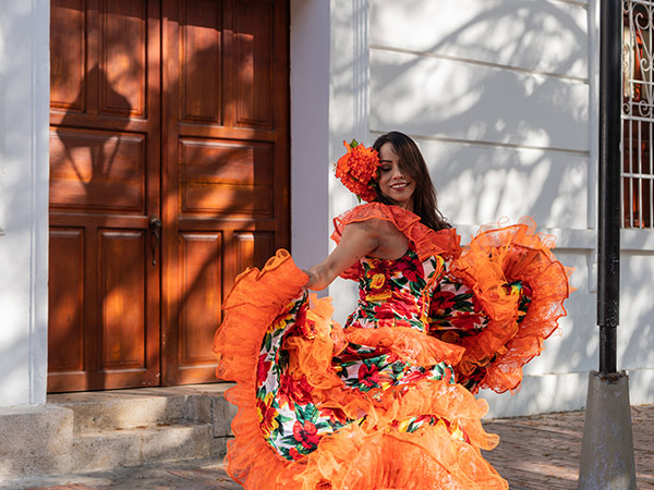 Smiling woman dancing in vibrant traditional dress outdoors against a wooden door on a sunny day in colombia