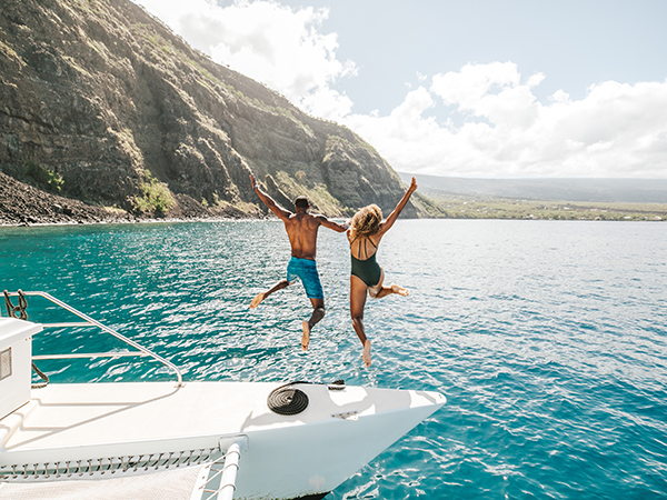 Two people jumping off a yacht with the beautiful coastline of Hawai&lsquo;i in the background.