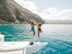 Two people jumping off a yacht with the beautiful coastline of Hawai‘i in the background.