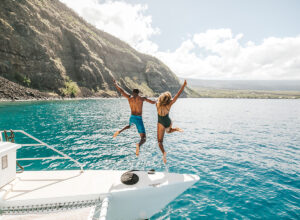 Two people jumping off a yacht with the beautiful coastline of Hawai‘i in the background.
