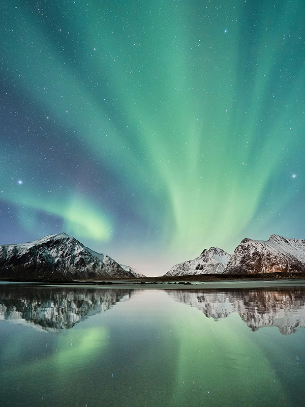 The aurora borealis dances across the night sky, illuminating rugged mountains and the calm, glistening sea below.