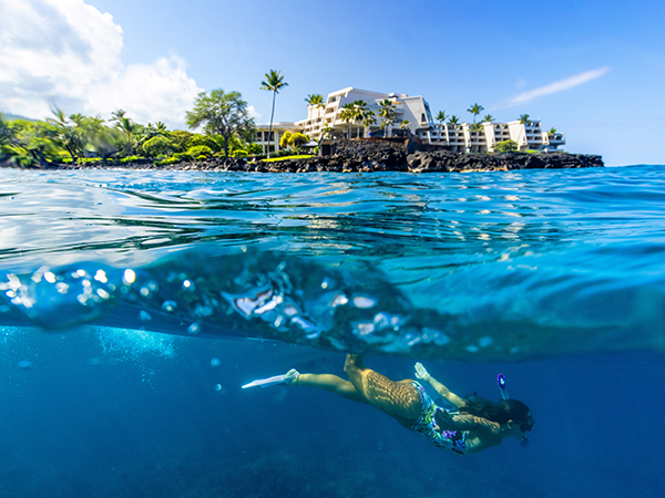 A woman diving beneath the sea, with the OUTRIGGER Hotel standing above the turquoise waters.